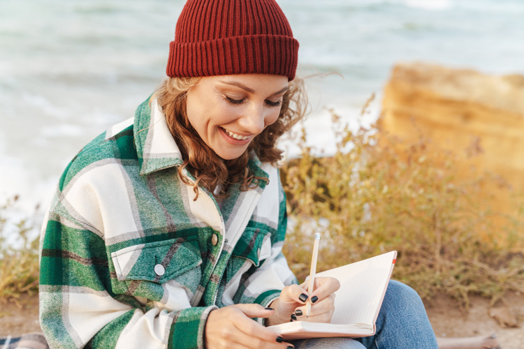Young woman in plaid shirt writes goals in her journal outdoors.